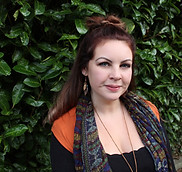 Woman with brown hair - Kelly Howard - and orange shirt smiling in front of green leaves.