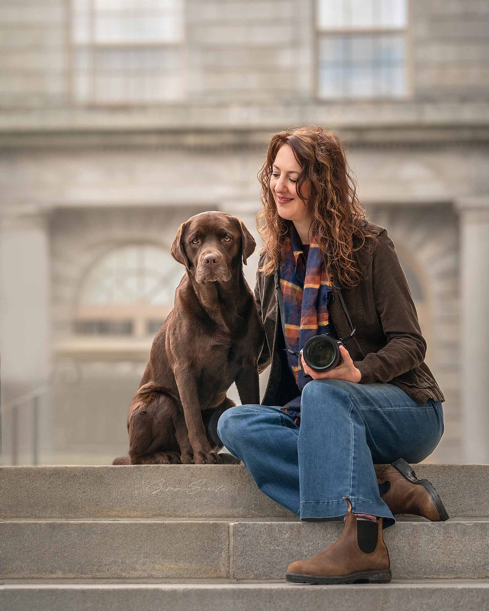 Jenny with curly hair sits on steps beside a chocolate Labrador, holding a camera. She's smiling. Background of a stone building.