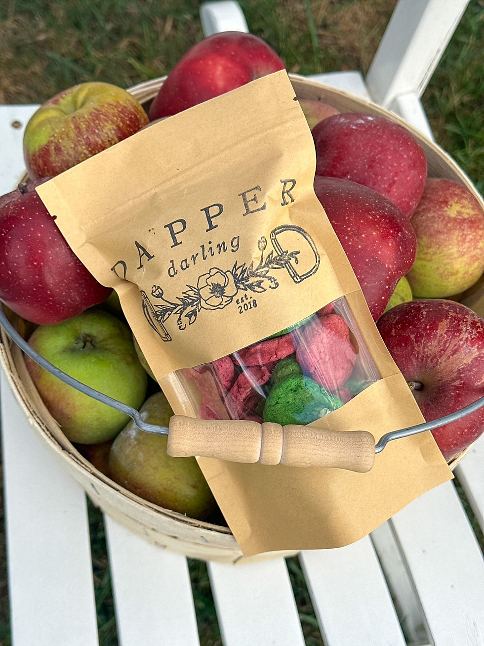 Basket of red and green apples on a white chair with a paper bag labeled "Dapper Darling" containing colorful snacks, outdoors.