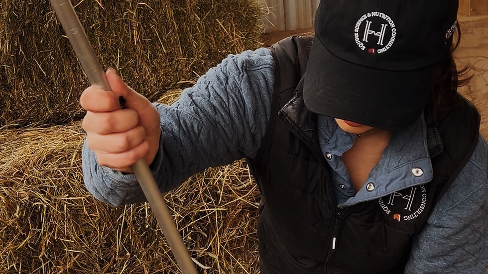 Person in blue jacket and black cap, holding a rake near hay bales. Cap reads "Equine Science & Nutrition Consulting." Rustic setting. Photo by Claudia D’Costa