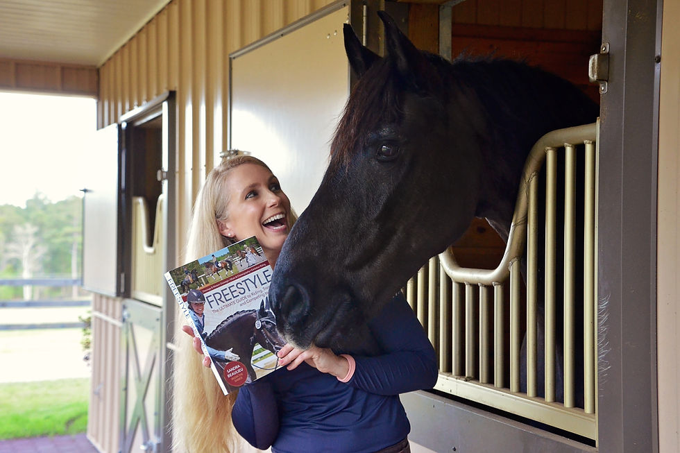 Smiling woman in a stable hugs a black horse and holds a Freestyle magazine; horse leans out of stall, playful and warm.