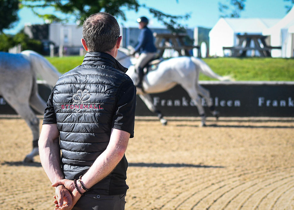 Man in Stonehall vest watches riders on white horses in an outdoor arena, with tents and picnic tables in the sunny background.