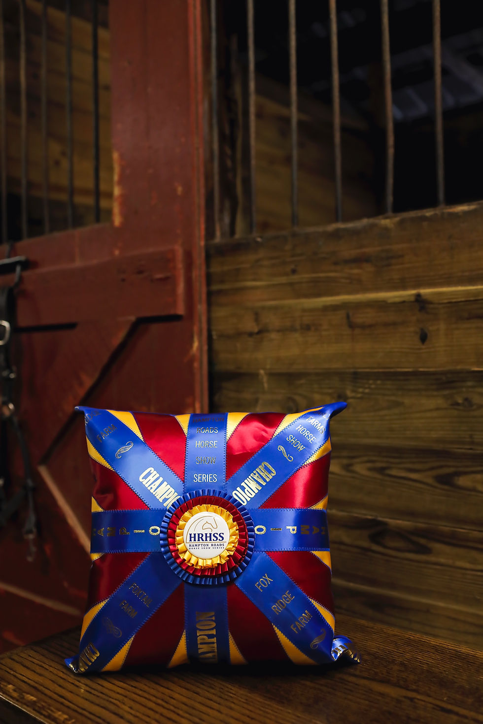Champion ribbon pillow with HRHSS Hampton Roads Horse Show Series text on a wooden bench in a rustic barn.