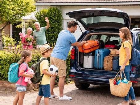 Familie packt vor dem Haus einen Kombi für den Urlaub. Die Nachbarn winken aus dem Garten, während Koffer und Taschen in den offenen Kofferraum geladen werden