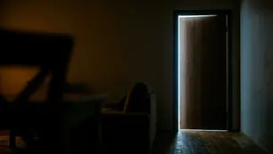 Blick aus einer dunklen Wohnung auf eine einen Spalt geöffnete Eingangstür mit kaltem Licht aus dem Treppenhaus. Das Bild zeigt die Situation, in der ein Sperrbügel oder eine Türkette die Tür sichert.