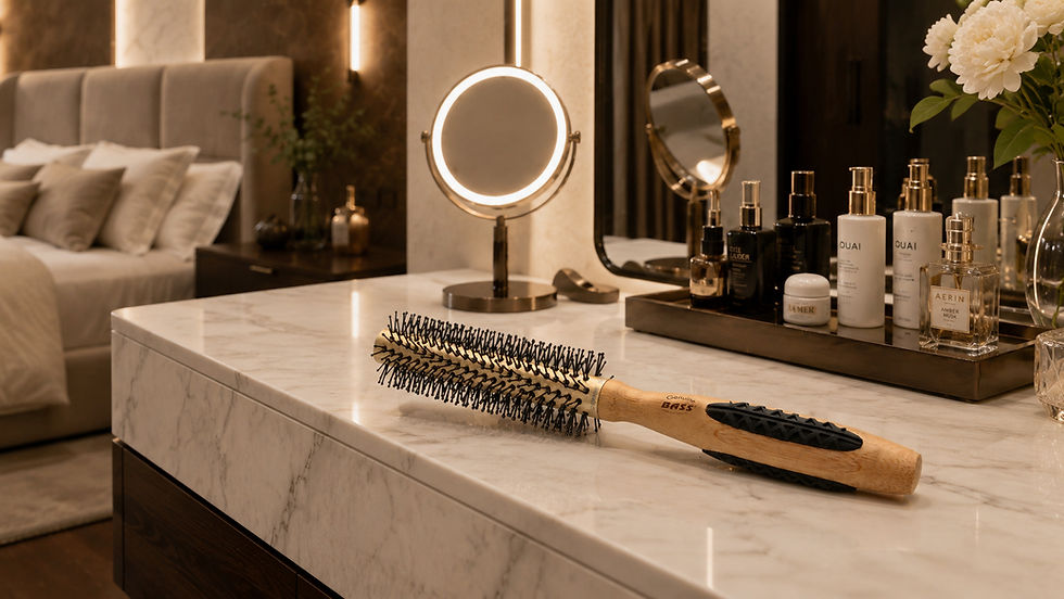 Bass Brushes Hairbrush on marble vanity in a warm bedroom, beside a lit round mirror, hair products, and white flowers; bed in background.