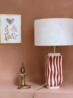 La lampe est posé sur un fond saumon avec en arrière plan un tableau, et sur la gauche il y a un objet pour habillé un peu plus la lampe. 