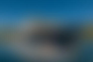 Couple rides jet ski past a large yacht in sunny, blue sea. The yacht is navy with cream details. Forested hills are in the background.