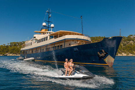 Couple rides jet ski past a large yacht in sunny, blue sea. The yacht is navy with cream details. Forested hills are in the background.