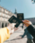 Two hands holding U.S. passports against a sunny plaza backdrop with historic buildings. The mood is travel-ready and excited.