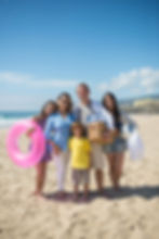 Family of five smiling on a beach. They hold a pink float, picnic basket, soccer ball, and towel. Bright sunny day with blue skies.