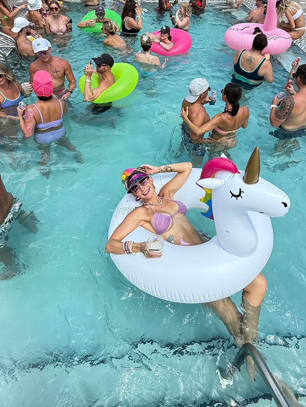 Woman in a unicorn float smiles, holding a drink in a crowded pool. She's in a purple bikini and colorful cap. Fun, relaxed atmosphere.