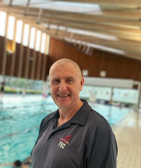 Coach Paul, a white man with no hair is smiling by the pool, wearing a dark Formby Swim Club polo shirt