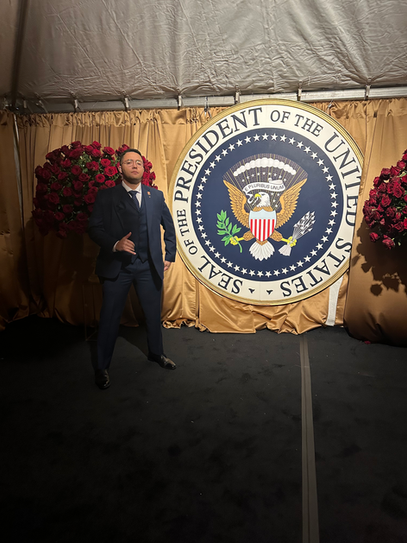 Man in suit standing in front of presidential seal backdrop at formal event