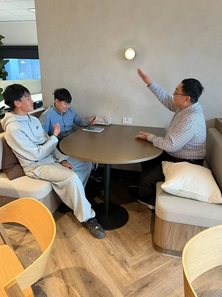 Three people sit at a round table in a modern room. One gestures energetically while others listen attentively. Light tones and wooden floor.