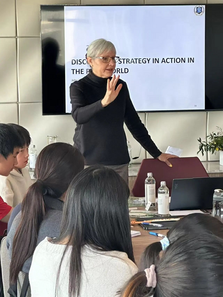 Woman presenting to a group in a classroom setting, gesturing with hand. “DISCOVERY STRATEGY IN ACTION” text on screen. Attentive audience.