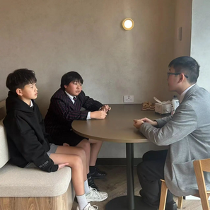 Three people sit around a table in a minimal room. Two children in school uniforms and an adult in a suit engage in conversation.