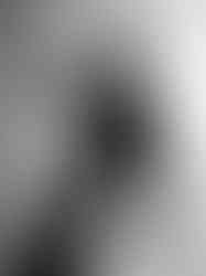 Black-and-white portrait of a man in a black turtleneck and jeans leaning back with one hand raised near his forehead against a plain gradient background.