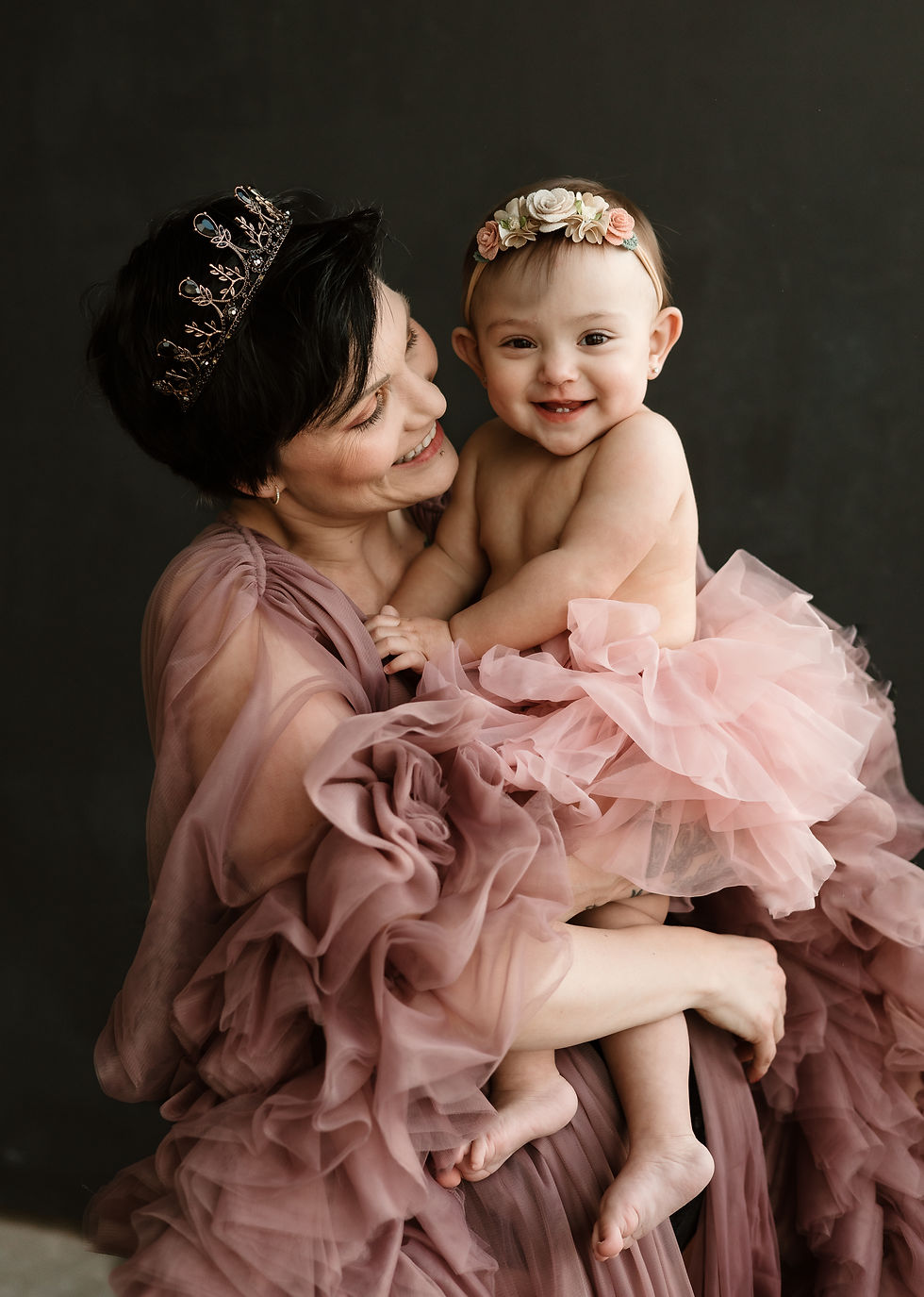 Woman in mauve dress holds smiling baby in pink tutu; both wear floral headbands. Dark background, joyful mood.