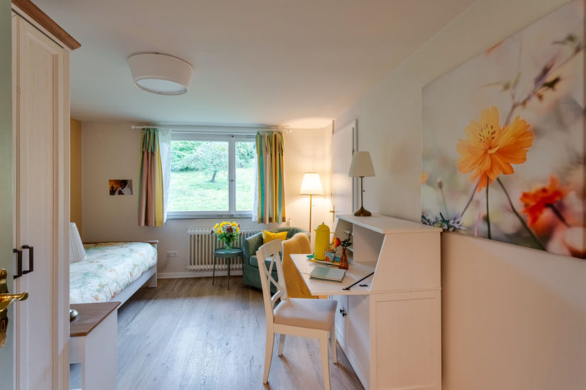 Helles, in Gelbtönen gehaltenes Zimmer mit einem schönen Schreibtisch rechtd uns einer Fotografie einer Kosmea. Links ein Einzelbett und gerade aus eine Fenster mit Blick auf Apfelblüten.