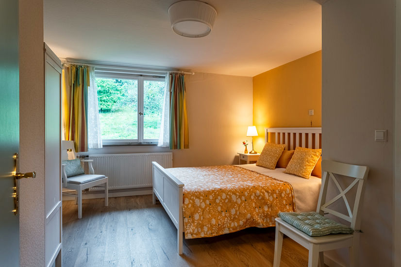 In Gelbtönen gehaltenes Doppelbettzimmer mit dem Blick aus dem Fenster auf Apfelbäume. Tür mit goldener Türklinke.