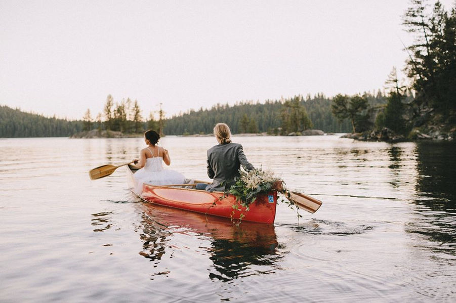 Wedding couple in red canoe decorated with flowers rowing away from camp.
