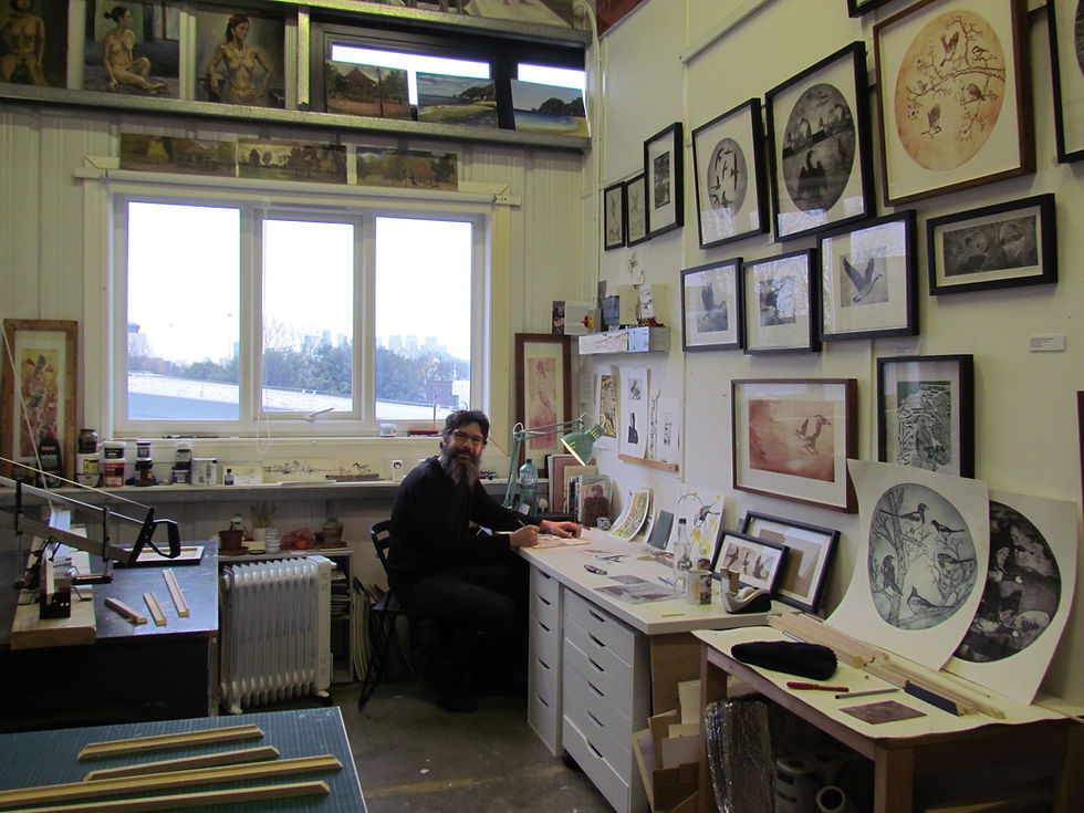 Artist Tim Mitchell sits in his studio at a desk, surrounded by his prints and framed pictures on the wall. Canary Wharf is in the background through a window.