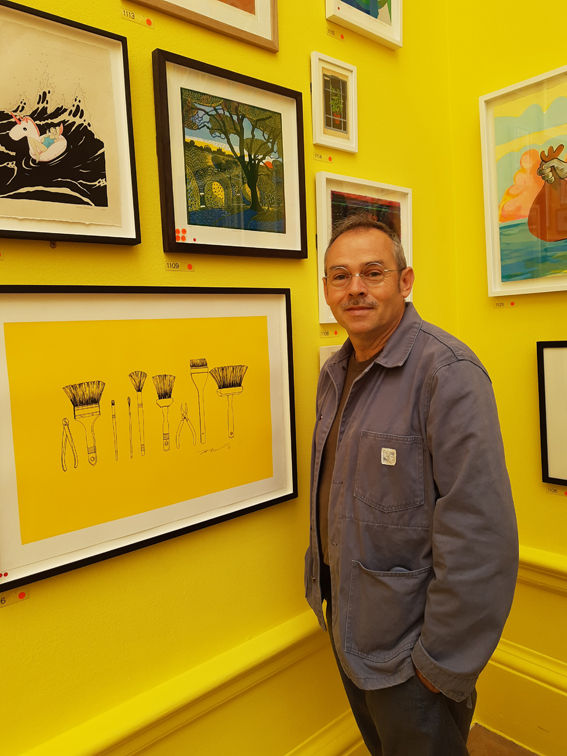 Artist Kit Boyd stands next to his framed Greenwich Park linocut which is hung on a bright yellow wall, surrounded by other pictures. There are red and orange dots on the frame, signifying sales.