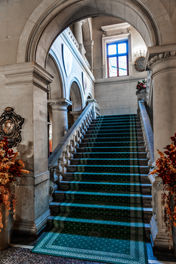 Monumentalne jasne schody z zielonym dywanem, klasyczną balustradą i ciemnym kasetonowym sufitem.