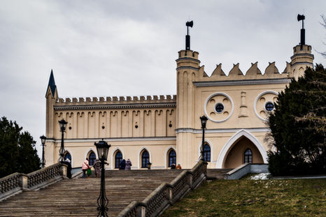 Neogotycka fasada Zamku Lubelskiego z rozetami, ostrołukowymi oknami i blankowaną attyką, z monumentalnymi schodami prowadzącymi do centralnego wejścia flankowanego wieżyczkami.