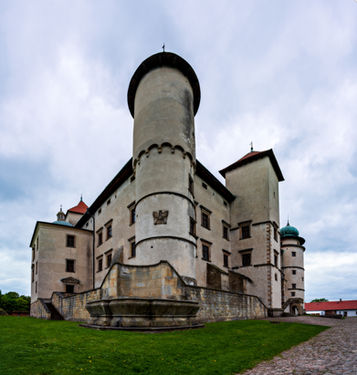 Widok z niskiej perspektywy na dziedziniec z wysoką cylindryczną wieżą na kamiennym cokole oraz skrzydłami z jasnymi elewacjami i wieżą z zieloną kopułą w tle pod pochmurnym niebem.