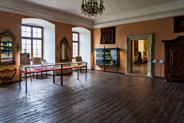 Przestronna galeria z drewnianą podłogą z szerokich desek, ochrowymi ścianami, wysokimi oknami po lewej, szklanymi gablotami z drobnymi przedmiotami, złoconymi lustrami i obrazami na ścianach, kryształowym żyrandolem oraz kamiennym portalem drzwiowym prowadzącym do kolejnej sali.