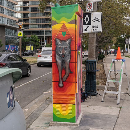 A grey coyote pads down a railway track. Around the coyote stretch bands of colour from red through orange, yellow, and green. A green heart hovers above the coyote's head.