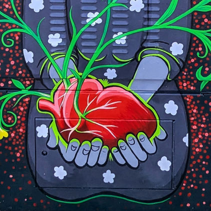 Close up of a woman's hands holding a large human heart -- she is kneeling in a floral dress in grey. The veins in the heart sprout into the roots of green flowering vines that stretch across the box. The dark background is peppered with red and pink dots.