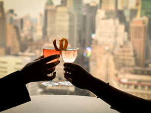 Silhouette of 2 arms, each with a cocktail overlooking a window of the NYC skyline