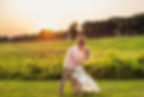 A couple kisses in a grassy field at sunset. The woman wears a floral dress, the man a pink shirt. Trees and a monument in the background. Romantic mood.