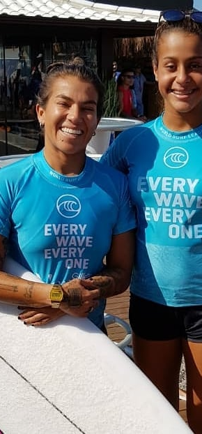 Surfista com prancha branca e outra garota ao lado , vestindo camisa azul