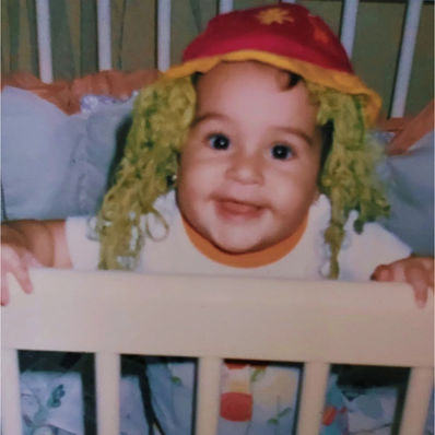bebê em pé no berço de chapéu com cabelo verde de palhaço, sorrindo