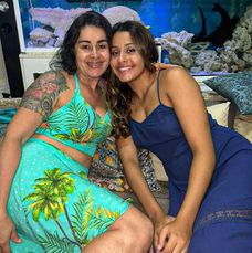 Mãe e filhas de rostos colados , uma do lado da outra a filha de vestido azul e a mãe de saia e top verde com detalhes de coqueiros, ambas sorrindo.