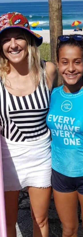 2 garotas uma de camisa azul e short preto e a outra de maio listrado brancoe preto de short branco segurando a prancha rosa e de chapeu vermelho na cabeça.