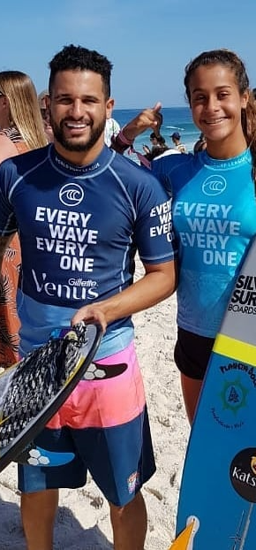Uma garota de camisa azul com prancha cinza com azul e o rapaz com camisa azul escura em um evento de surf.