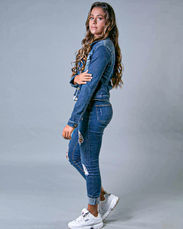 Garota de calça e jaquetas jeans, de cabelos compridos segurando o braço com a mão direita e o braço esquerdo reto sobre o corpo de tenis sem meia  branco de lado em um fundo cinza.