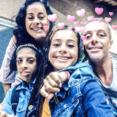 Familia em ordem de tamanho menina menor na frente, a outra irmã atrás e seguidos de pai e mãe, todos abraçados.