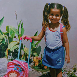 Menina com cabelos presos  de chuquinhas com saia azul e blusa cinza e de mochila de rodas da moranguinho.