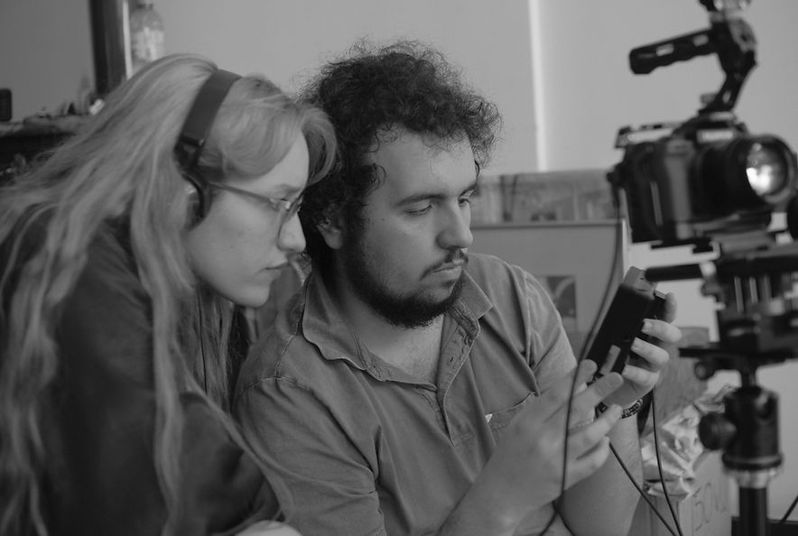 Fotografía en blanco y negro de un hombre y una mujer jóvenes observando una cámara. Ella lleva auriculares.