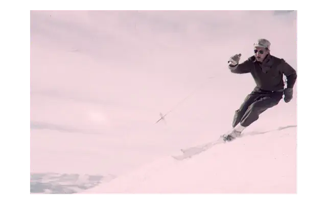 Man skiing downhill on snowy slope, wearing dark clothing and sunglasses, with overcast sky in the background. Dynamic motion and focus.