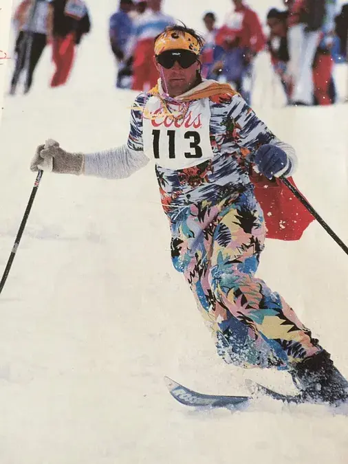 Skier in colorful outfit with "Coors" bib 113, skiing downhill on snowy slope, surrounded by people in the background, displaying energy.