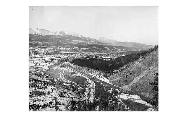 Snow-covered valley with winding river and trees, surrounded by distant mountains under a clear sky. Serene, vast natural landscape.