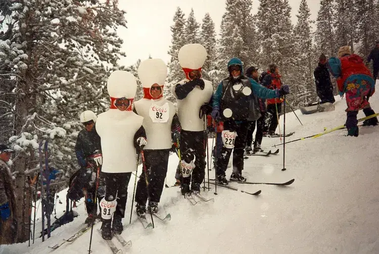 Skiers in snowman costumes with race numbers stand on a snowy slope, surrounded by snow-covered trees. They appear joyful and playful.