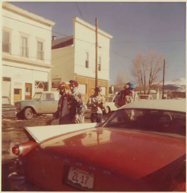 Clowns walk past a parked red Dodge in a small town with old buildings. The sky is clear, and mountains are visible in the background.